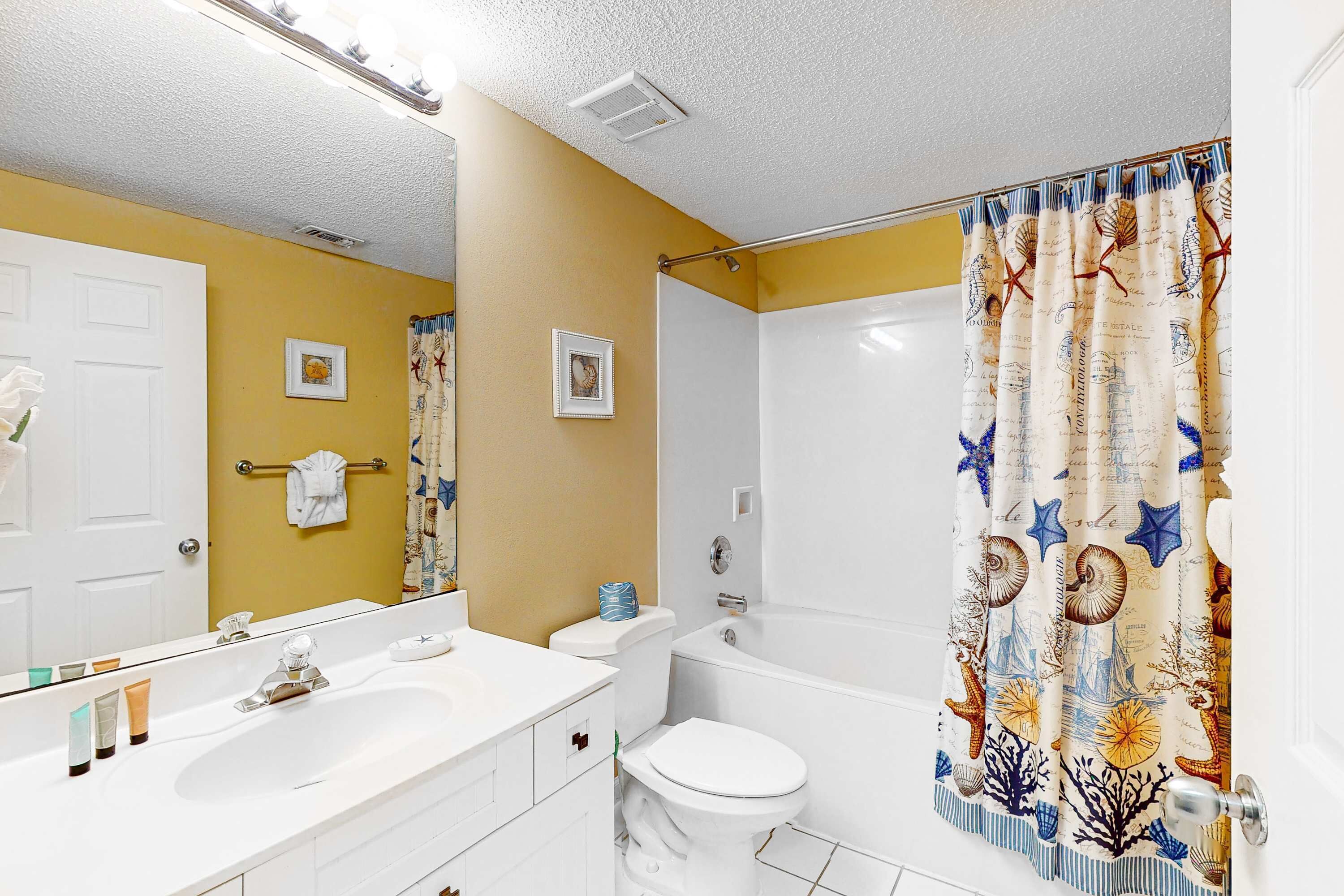 Une salle de bain avec un lavabo, des toilettes et une baignoire, tous de couleur blanche. Un miroir est placé au-dessus du lavabo, et un porte-serviettes est fixé au mur.