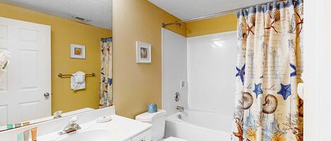 Une salle de bain avec un lavabo, des toilettes et une baignoire, tous de couleur blanche. Un miroir est placé au-dessus du lavabo, et un porte-serviettes est fixé au mur.