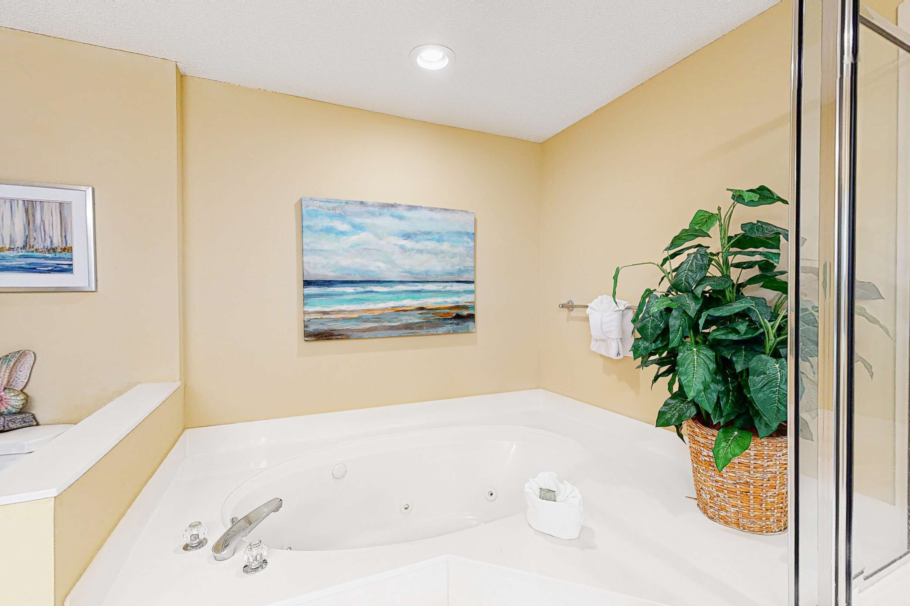 A bathroom with a large bathtub, a painting of a beach scene, a potted plant, and a glass shower enclosure.