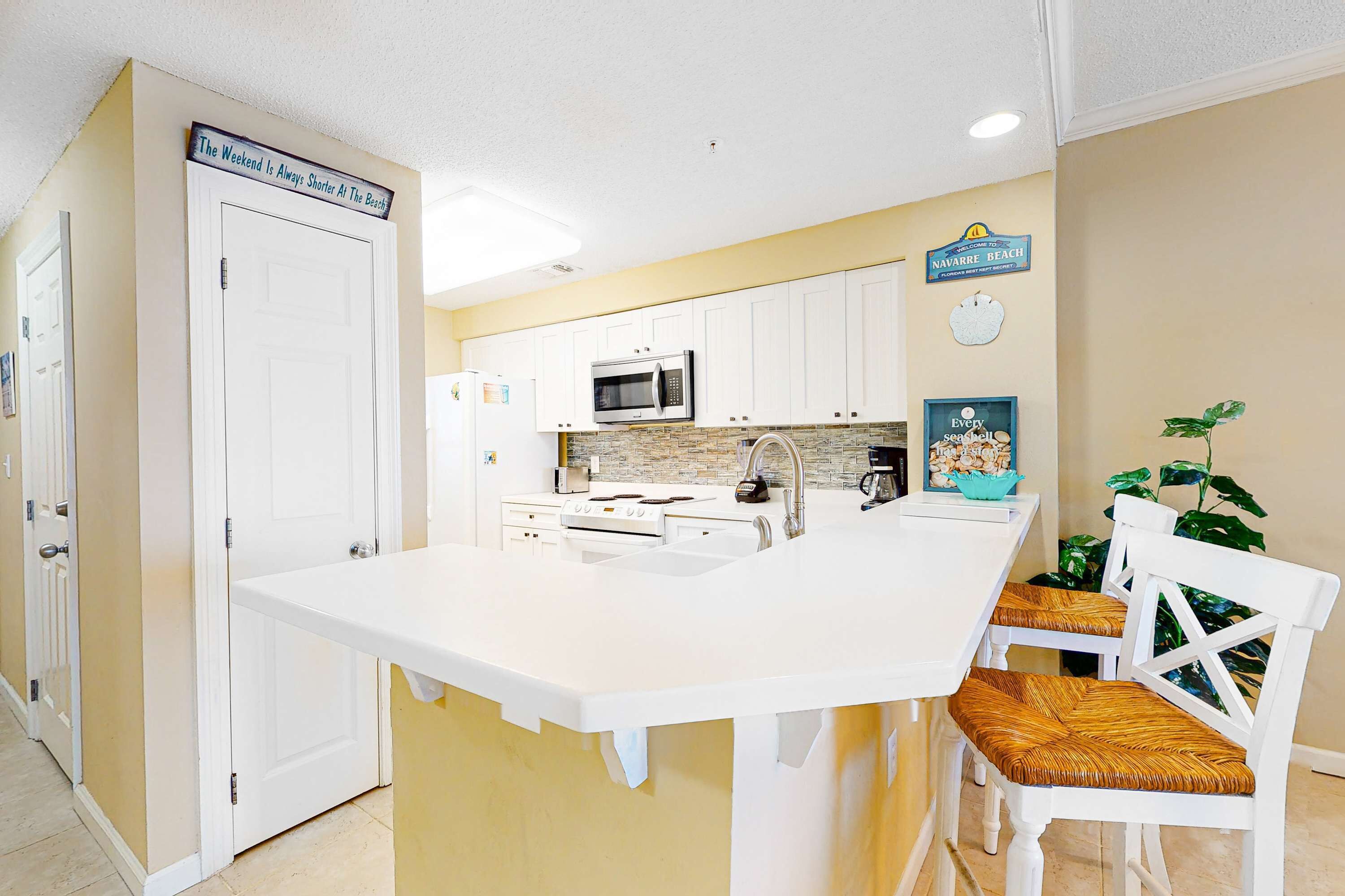 A kitchen with white cabinets, a microwave, and a sink. There is a sign on the wall that says "The Weekend Is Always Shorter At The Beach."