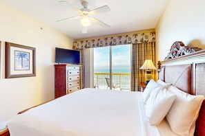 Een hotelkamer met een bed, een televisie, een plafondventilator, een raam met uitzicht op het strand en een balkon met een stoel.