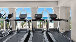 Un gimnasio con cintas de correr y equipo de ejercicio, ventanas amplias con vistas a la playa y palmeras.