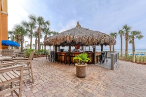 A beachside bar area with wooden chairs and tables, a thatched roof, and palm trees. - Open & spacious condo in beachfront complex with pool, gym, elevator & fast WiFi (Panama City Beach)