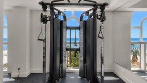Un gimnasio con barra de dominadas y máquinas de pesas, ventanas amplias con vista a la playa.