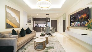 Sala de estar moderna com sofá, mesa de centro e área de jantar. Uma cozinha aparece ao fundo e uma televisão grande na parede.