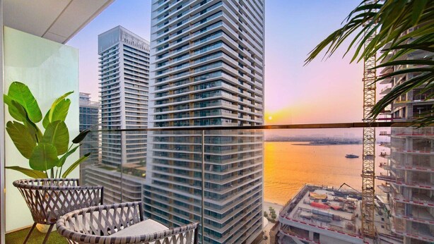 Um edifício alto com vista para o pôr do sol sobre a água, uma varanda com mobiliário de vime e uma grua de construção ao fundo.