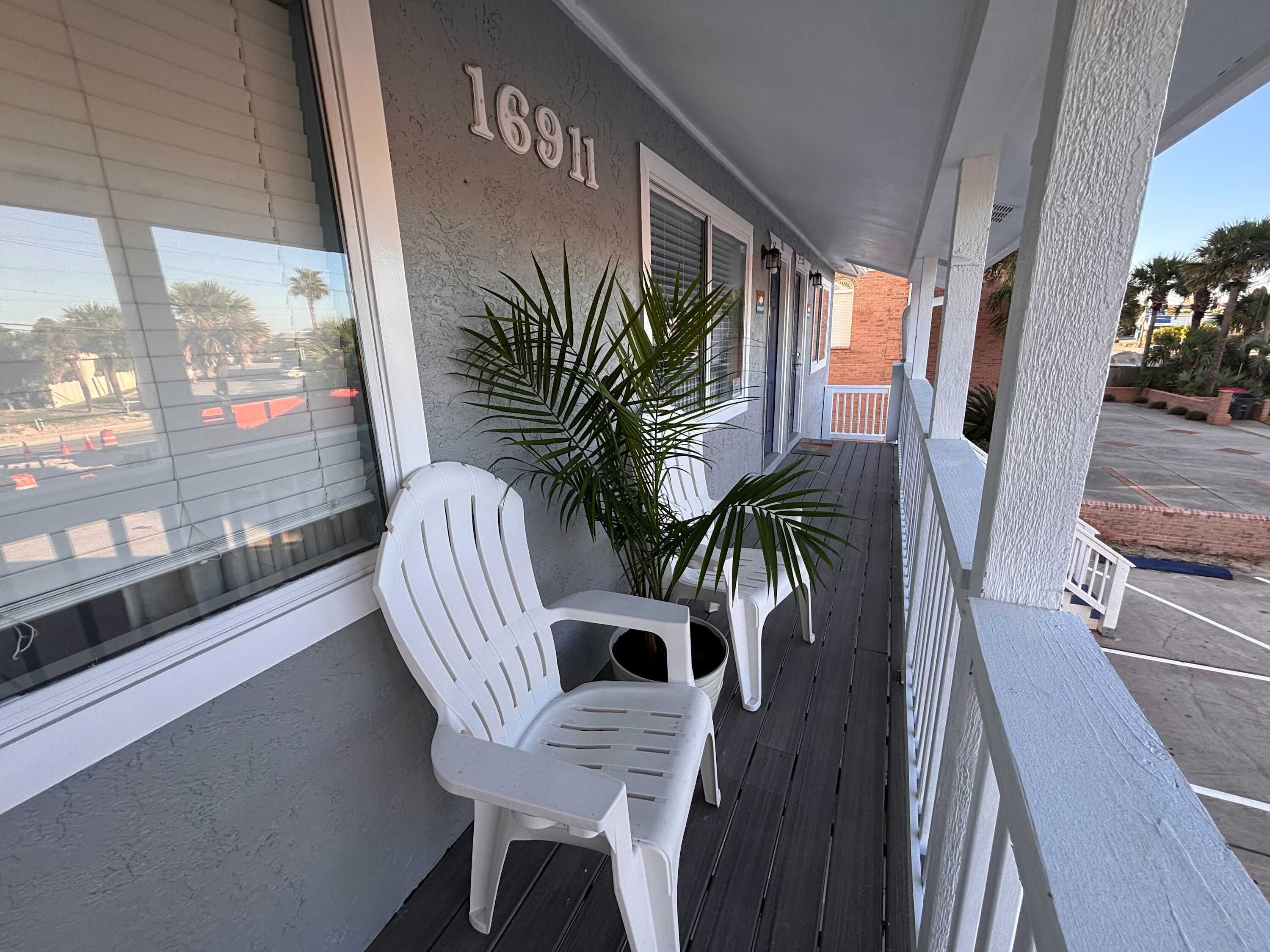 A covered porch with white chairs and a potted plant, a number '1691' on the wall, and a view of the parking lot.