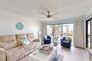 A living room with a beige sofa, a coffee table with a woven basket, a ceiling fan, and a view through large windows. - Beachview Condo with full kitchen, shared pool, hot tub and beach access (Panama City Beach)