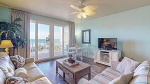 A living room with a sofa, coffee table, and TV, a balcony with a view, and a ceiling fan.