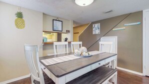 A dining area with a dark wooden table, white chairs, and a pineapple decoration on the wall. - Bright condo in gated community with pool, hot tub & tennis (Panama City Beach)