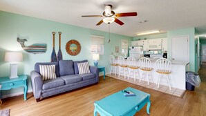 A living room with a sofa, a coffee table, a ceiling fan, and a kitchen area with white cabinets and a microwave. - Beachfront condo with true beachfront walkout, patio, BBQ area, & Gulf view (Panama City Beach)
