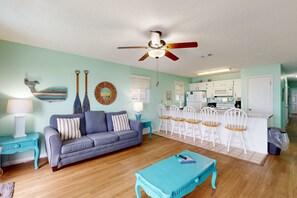 A living room with a sofa, a coffee table, a ceiling fan, and a kitchen area with white cabinets and a microwave. - Beachfront condo with true beachfront walkout, patio, BBQ area, & Gulf view (Panama City Beach)