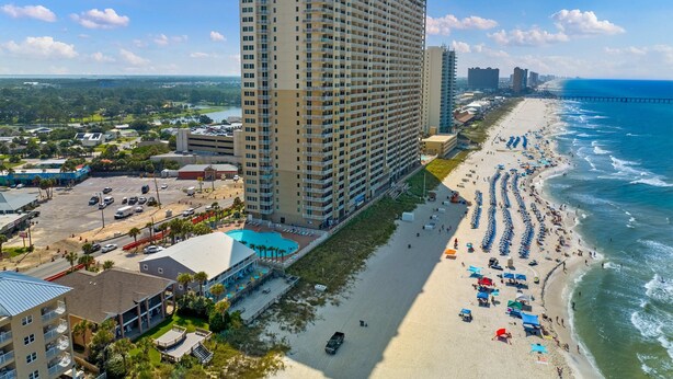A high-rise hotel with a beach view, a parking lot, and a long stretch of beach with umbrellas and people. - Beachfront condo with true beachfront walkout, patio, BBQ area, & Gulf view (Panama City Beach)