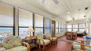 Ein Zimmer mit Korbmöbeln, Deckenventilator und großen Fenstern mit Jalousien.