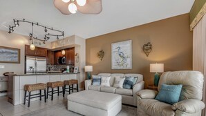 Ein modernes Wohnzimmer mit einer Couch, einem Hocker und Barhocker. Es gibt eine Küchenzeile mit Kühlschrank und Mikrowelle.