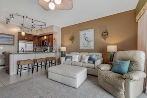 A modern living room with a sofa, ottoman, and bar stools. There is a kitchen area with a refrigerator and microwave. - Beachfront condo with Gulf views, pools, hot tub & free beach chair service (Panama City Beach)