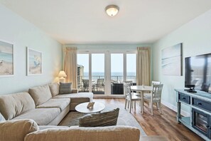 Ein Wohnzimmer mit Sofa, Couchtisch und Fernseher, ein Balkon mit Meerblick sowie ein Essbereich mit Stühlen und Tisch.