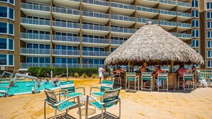 Un área junto a la piscina con sillones de descanso y un bar, rodeada por un edificio de varios pisos con balcones.