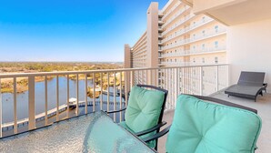 A balcony with a glass table and two green cushioned chairs, overlooking a body of water and a building with multiple balconies. - Water View Condo with Balcony, Central AC, WiFi, Pool/Hot Tub/Gym - Snowbirds OK (Panama City Beach)
