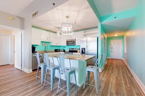A modern kitchen with white cabinets, a central island with bar stools, a stainless steel refrigerator, and a microwave. - Water View Condo with Balcony, Central AC, WiFi, Pool/Hot Tub/Gym - Snowbirds OK (Panama City Beach)
