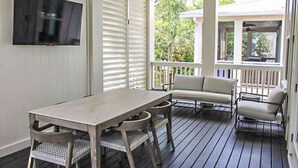 A covered porch with a wooden table and chairs, a television mounted on the wall, and a view of greenery outside. - Modern beachside haven with LSV and gas grill - bike to Blue Mt Beach (Santa Rosa Beach)