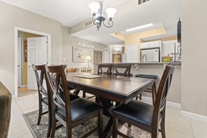 A dining area with a rectangular table and six chairs, a chandelier, and a kitchen in the background. - 7th Floor Lovely Condo w/ Outdoor Pool Heated Seasonally, On The Beach (Navarre Beach)