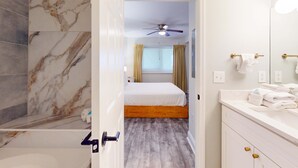 Bagno d'hotel con parete doccia in marmo, lavandino bianco con maniglie dorate e ventilatore a soffitto.