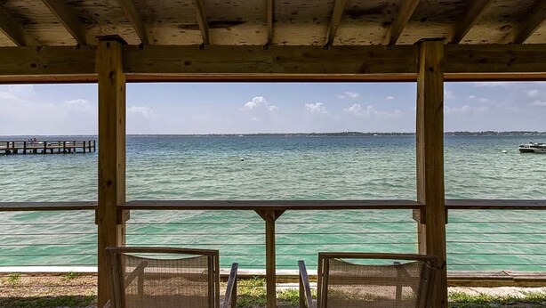 Un pontile di legno con due sedie rivolte verso un mare tranquillo, un molo sullo sfondo e un cielo sereno.