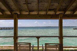 A wooden deck with two chairs facing a calm sea, a pier in the background, and a clear sky.