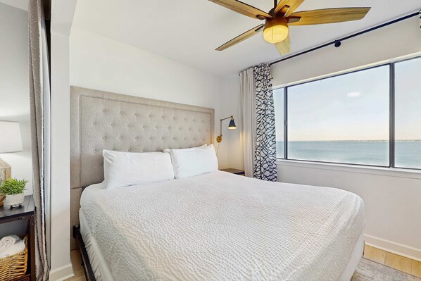 A bedroom with a large bed, a ceiling fan, a nightstand, and a window with a view of the sea.