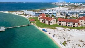 Vista aerea di un hotel costiero con diversi edifici, un molo che si protende nell'acqua e una spiaggia attrezzata con ombrelloni.