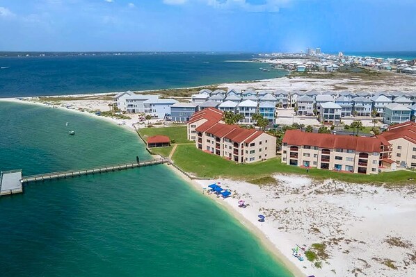 An aerial view of a coastal hotel with multiple buildings, a pier extending into the water, and a beach area with umbrellas. - Cozy coastal condo with water view, pool, central AC, & easy beach access (Navarre Beach)