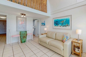 A spacious living area with a beige sofa, a glass coffee table, and a wooden side table with a lamp. There are two chairs with pink cushions and a decorative vase on the coffee table. - Dog-friendly beachfront condo with pool, hot tub, AC, & Gulf views (Panama City Beach)
