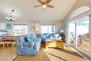 A spacious living room with a ceiling fan, a dining area with a wooden table and chairs, and large windows offering a view of the outdoors. - Gulf-front house near golf with pool, ocean views, beach access, & balcony (Inlet Beach)