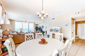 A modern living room with a round dining table, white chairs, a chandelier, and a view of the sea through large windows. - 11th Floor Gulf Front Condo Great Amenities & Private Beach Access (Navarre Beach)