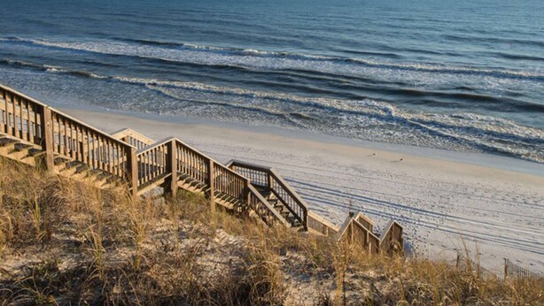 Un escalier en bois mène à une plage, avec l’océan en arrière-plan.