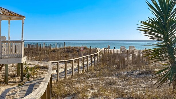 A wooden deck leading to a beach area with a wooden structure, possibly a gazebo, and a palm tree. - Gorgeous home with private pool, outdoor space and close to beach (Panama City Beach)