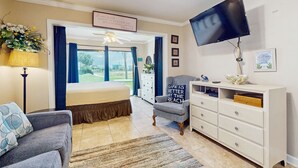 Een slaapkamer met een bed, een televisie, een bureau en een stoel. Er is een raam met gordijnen, een plafondventilator en een bord op de muur.