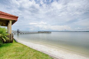 A beach with a wooden pier extending into calm waters, a covered area on the left, and a clear sky.