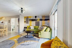 Una sala de estar moderna con un sofá verde, un sillón amarillo y una mesa de centro. Se ve una escalera que lleva a otra habitación y una puerta a la izquierda.