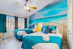 Habitación de hotel con dos camas, ropa de cama color turquesa, ventilador de techo y un ventanal con cortinas.