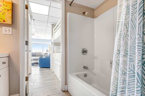 A modern bathroom with a bathtub and shower curtain, a white cabinet, and a view of a living area with a sofa and a blue chair. - Beachfront condo with central AC, WiFi, access to pool, washer&dryer (Panama City Beach)