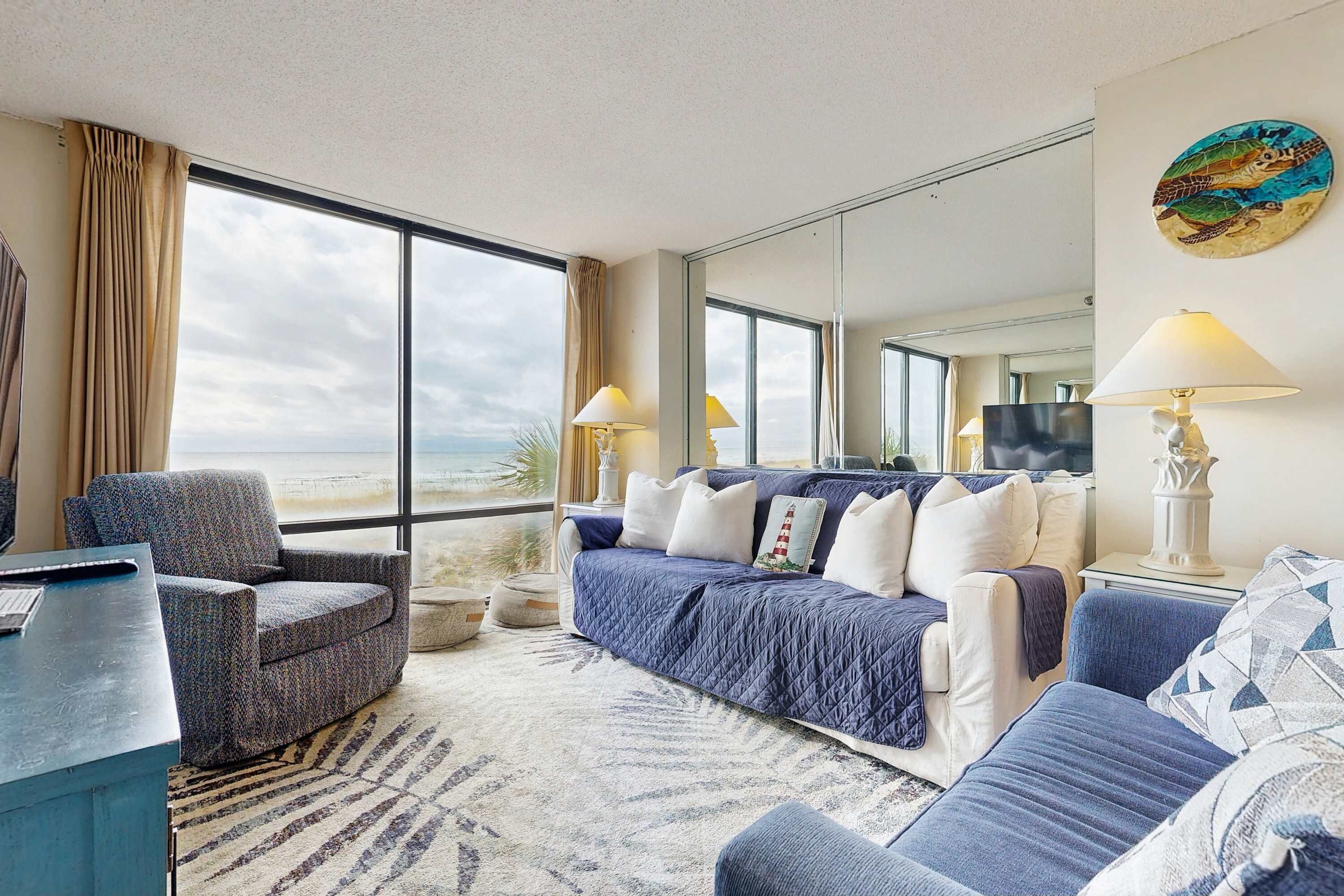 A modern hotel room with a large window offering a view of the beach, a comfortable seating area with a sofa and armchairs, and a flat-screen TV on a stand.