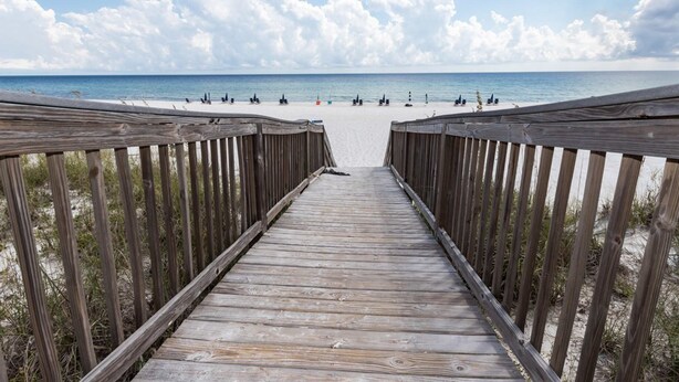 Un pont de fusta que condueix a una platja de sorra, amb una vista clara de l'oceà i un cel amb nuvolades.
