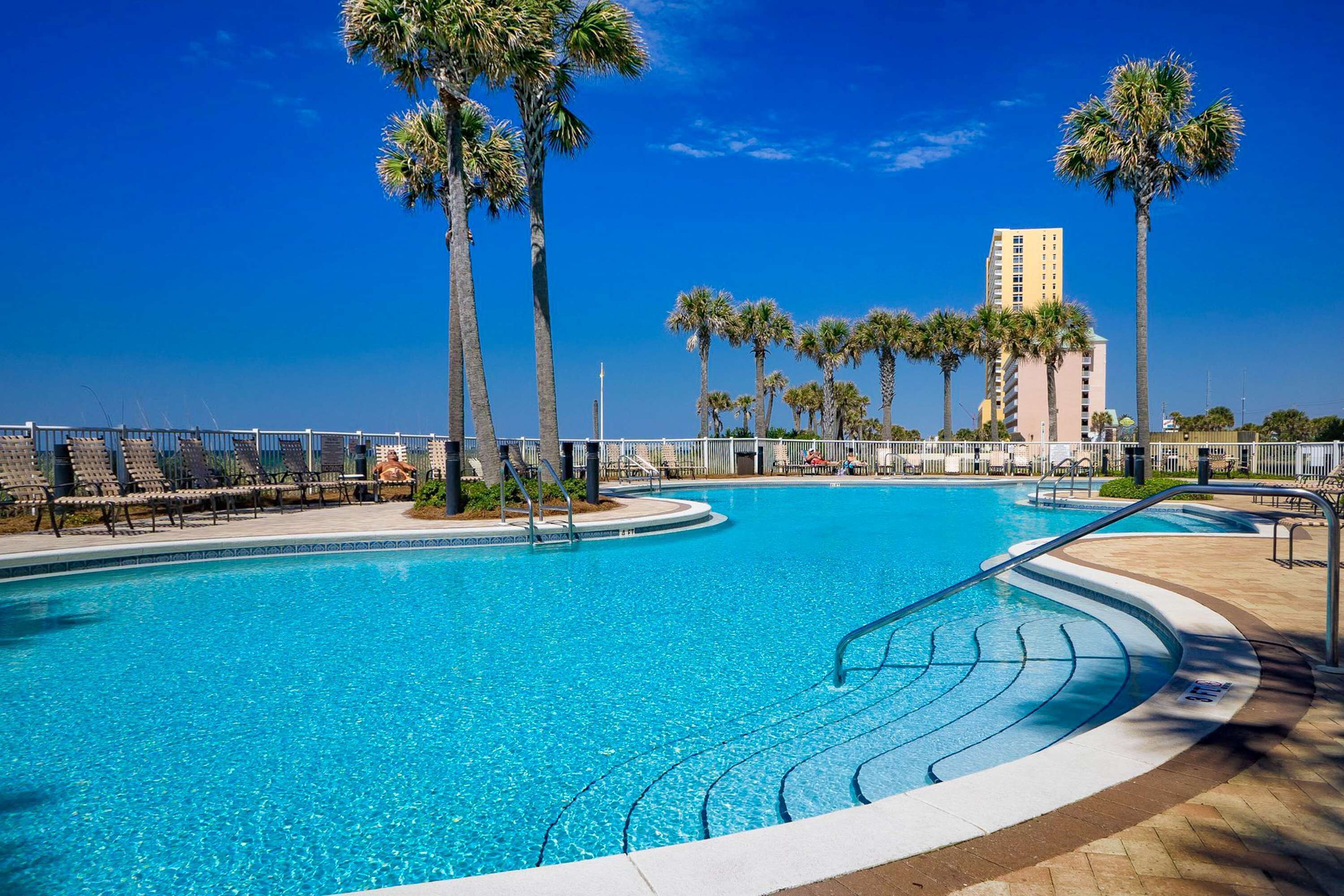 A swimming pool with palm trees, lounge chairs, and a tall building in the background.