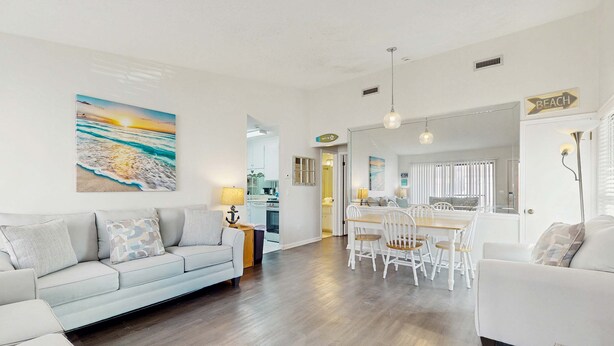 Ein geräumiger Wohnbereich mit einer Couch, einem Esstisch und Stühlen, einer Küche im Hintergrund und einem Wandbild im Strandhausstil.