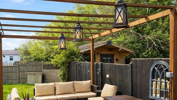 A covered outdoor patio with a wooden pergola, cushioned seating, and hanging lanterns. - Dreamy Stay With Cosy Charm (Addlestone)