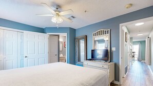 A bedroom with a large bed, a ceiling fan, a dresser, a TV, and a hallway leading to other rooms. - Bayview condo only steps from the beach w/ shared pool and peaceful location (Navarre Beach)