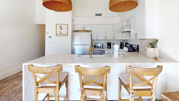 Una cocina moderna con armarios blancos, una isla central con taburetes y dos lámparas colgantes de mimbre.