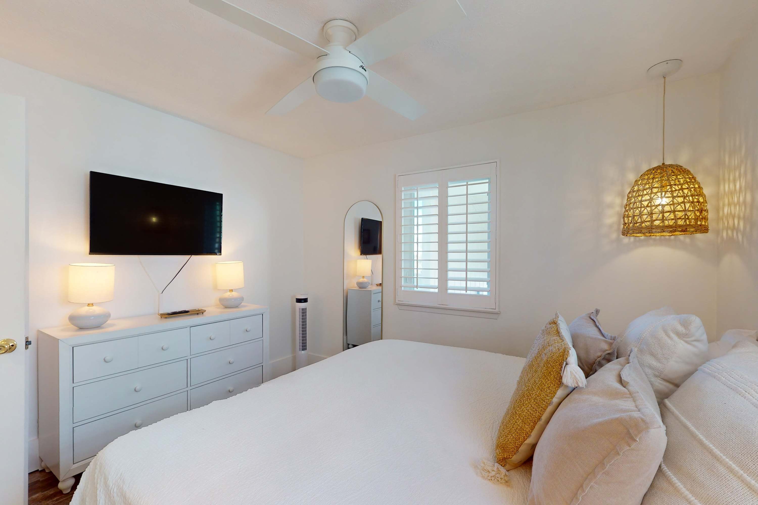 Een slaapkamer met een bed, een aan de muur bevestigde televisie, een plafondventilator en een raam met zonwering.
